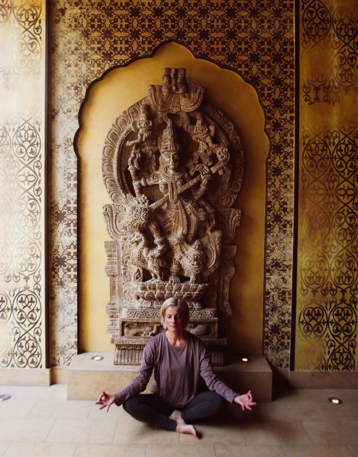 Korinna Schroll sitzt in Meditationshaltung vor einer goldenen Wand mit Steinrelief